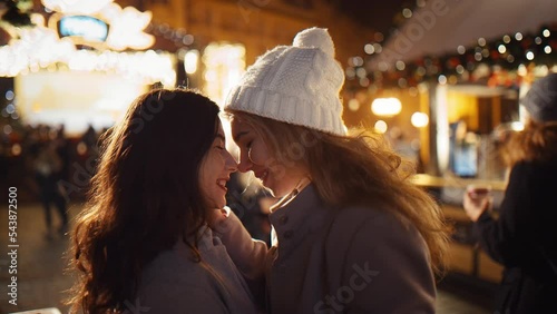 Two cheerful multi-racial girls hug and showing love, rubbing their noses in the middle of a busy New Year's Fair under the chimes of the New Year Christmas. Concept LGBT happiness freedom love photo