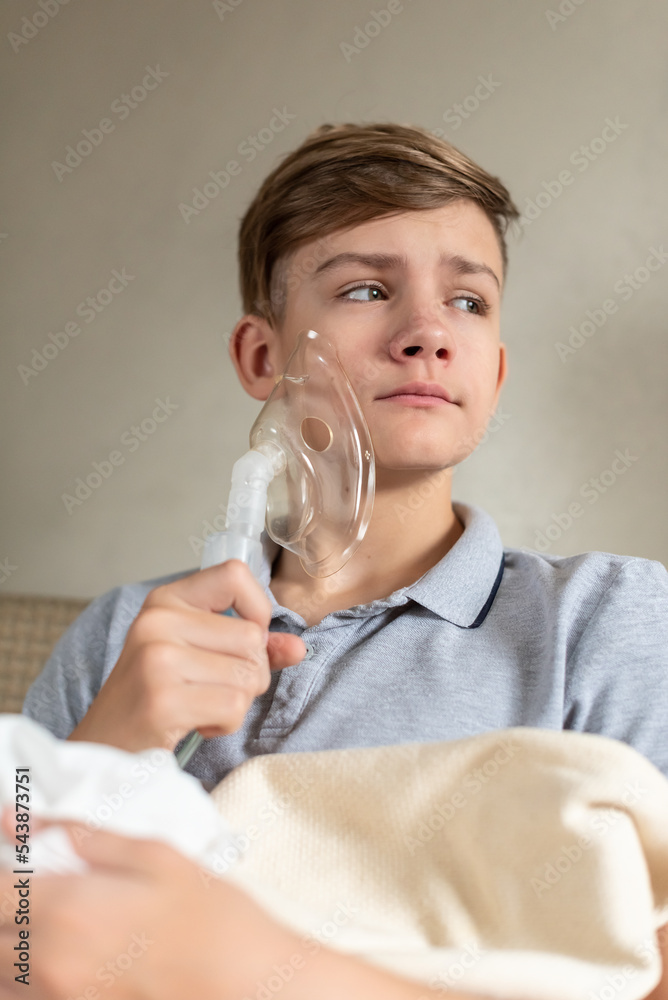 Sick teen boy with an inhaler. Unhealthy child doing inhalation at home, she use nebulizer and inhaler for the treatment, sitting on the couch at home. Asthma inhaler, nebulizer steam, flu or cold