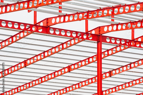 Fototapeta Naklejka Na Ścianę i Meble -  Red castellated beam metal roof structure of large industrial building in construction site against white cloudy sky background