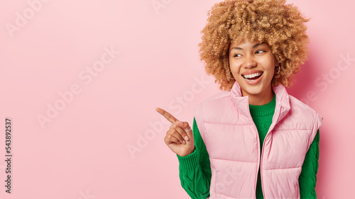 Photo of good looking woman with curly hair points index finger and shows advertisement smiles happily shows white teeth wears green lomg sleeved jumper and vest isolated on pink background. photo