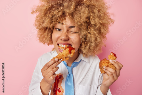 Photo of hungry young European woman eats delicious fried chicken nuggets with ketchup winks eye enjoys eating fatty appetizing unhealthy food dressed in formal clothes isolated over pink background