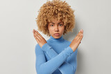 Serious strict woman crosses arms makes taboo gesture says enough or no demonstrates denial stands against grey studio background dressed in casual blue turtleneck wants to finish conversation