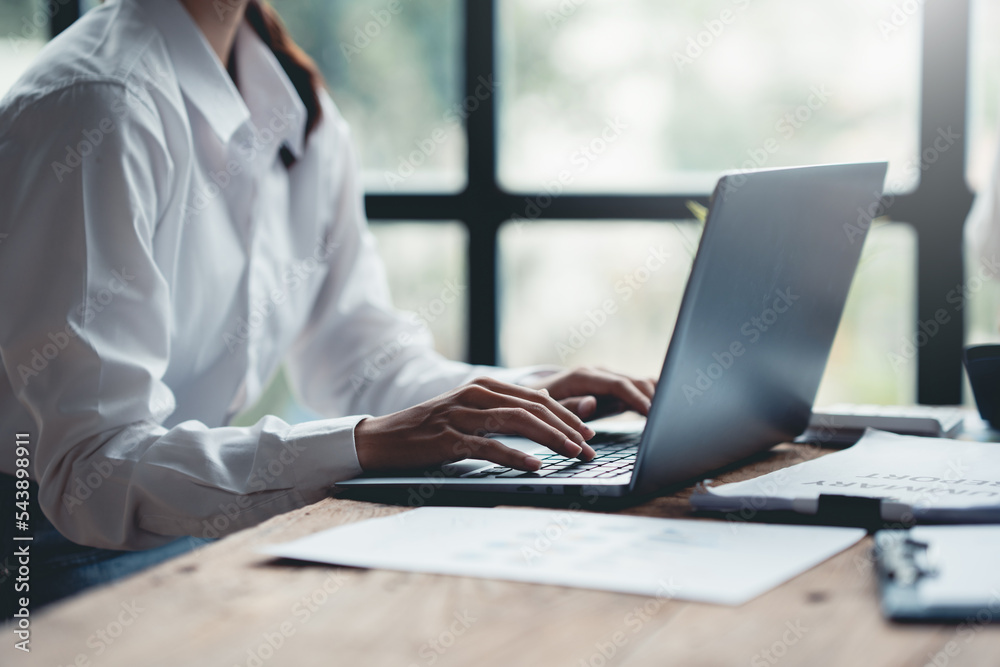 Close-up of the hands of a business woman attentively typing on a laptop computer and having a document on the desk in the office. Close-up of the hands of a business woman attentively typing on a laptop computer and having a document on the desk in the office.