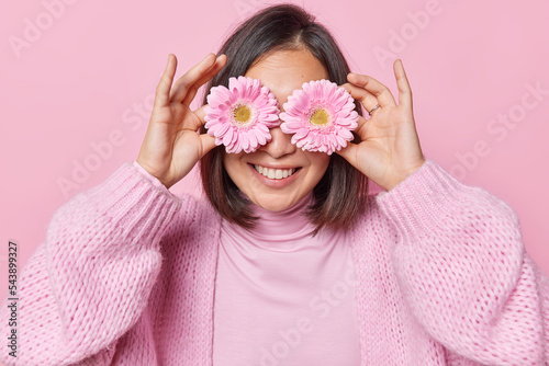 Horizontal shot of pleased unrecognizable dark haired woman covers eyes with gerbera flowers smiles toothily enjoys pleasant fragrant dressed in casual knitted jumper isolated over pink background