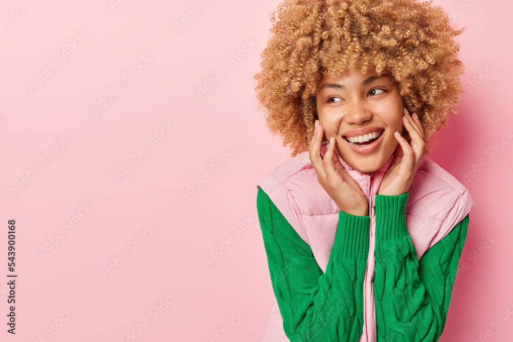Happy woman with curly hair smiles broadly keeps hands on cheeks has healthy skin white teeth dressed in green jumper and vest looks gladfully aside isolated over pink background blank space Happy woman with curly hair smiles broadly keeps hands on cheeks has healthy skin white teeth dressed in green jumper and vest looks gladfully aside isolated over pink background blank space