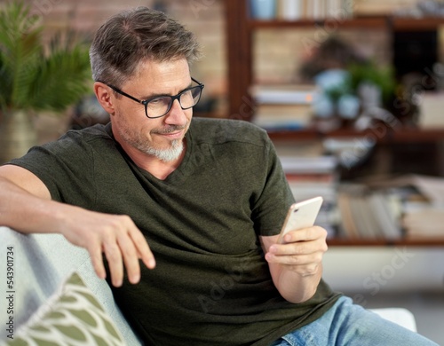 Portrait of middle aged handsome man wearing casual clothes, sitting on sofa at home in the living room, using phone.