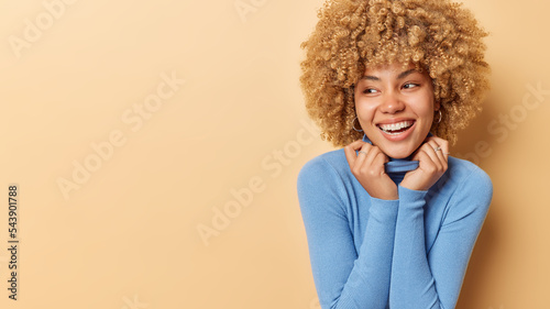 Indoor shot of sincere curly haired woman looks happy aside keeps hands on collar of turtleneck feels glad expresses positive emotions isolated over beige background copy space for your promotion