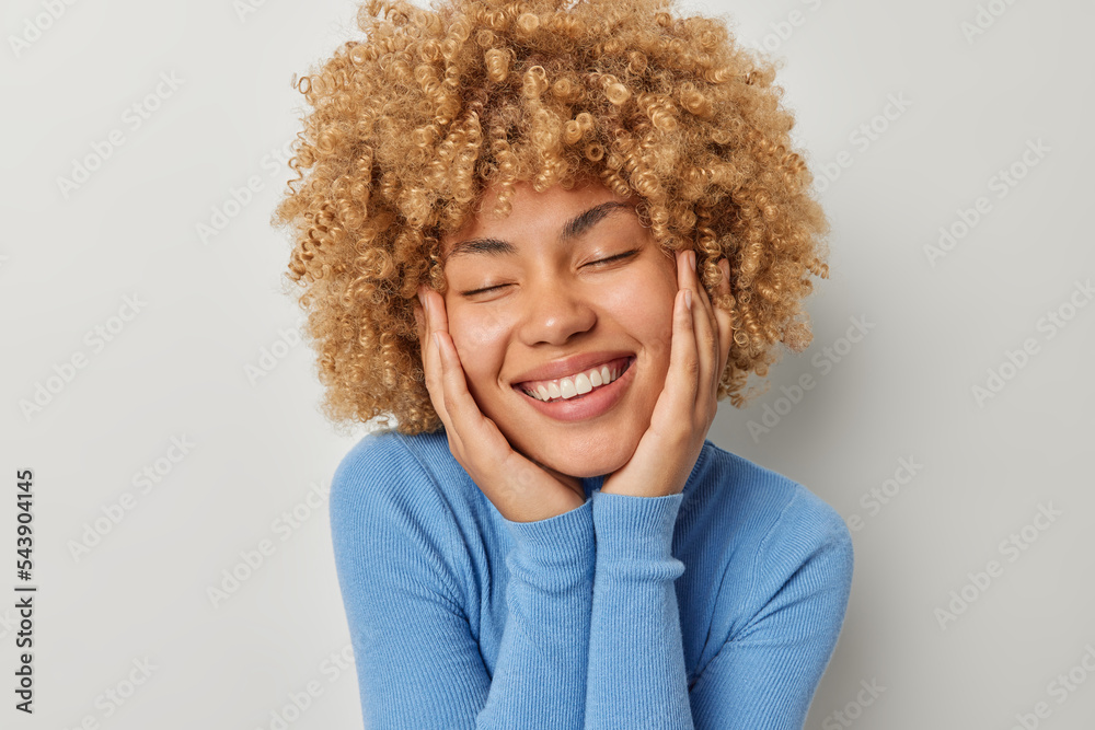 Portrait of cheerful curly haired woman keeps hands on cheeks stands with eyes closed smiles toothily recalls pleasant memories feels happy wears casual blue turtleneck isolated over grey background Portrait of cheerful curly haired woman keeps hands on cheeks stands with eyes closed smiles toothily recalls pleasant memories feels happy wears casual blue turtleneck isolated over grey background