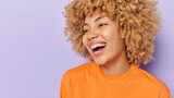 Horizontal shot of pretty cheerful woman with curly hair smiles broadly shows white teeth wears orange jumper expresses positive emotions isolated over purple background copy space for your text