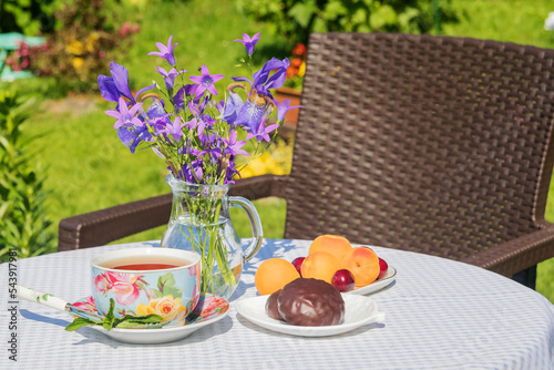 On the garden table, a cup of tea, chocolate marshmallows, fresh fruits. A bouquet of flowers in a vase.