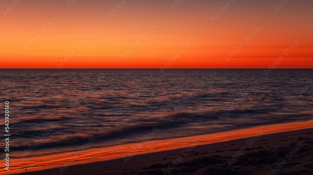 Fototapeta premium Vibrant orange red sky reflecting in calm ocean after sunset