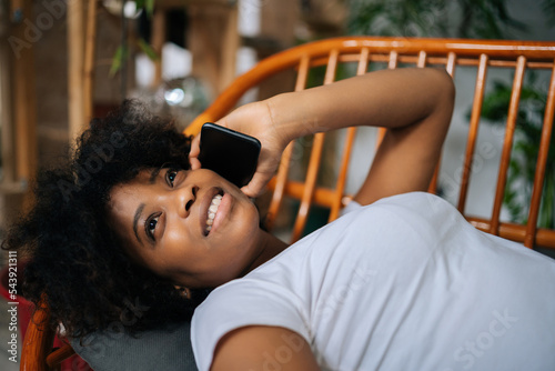Close-up high-angle view of cheerful African young woman talking on smartphone lying on comfortable couch having pleasant phone talk in living room at home, with modern green biophilic interior design