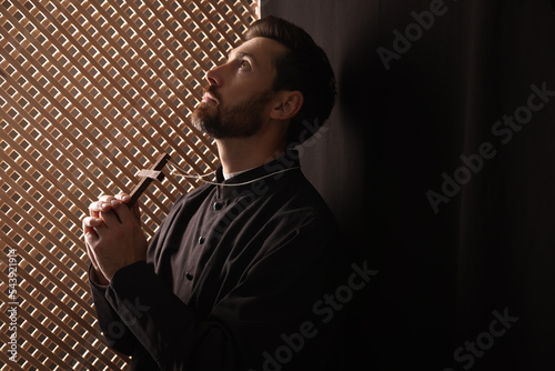 Фотография Catholic priest in cassock holding cross and praying to God in confessional, spa