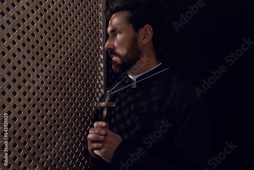Catholic priest in cassock holding cross in confessional booth, space for text photo