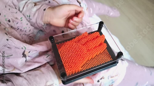 Young girl playing with plastic pin art toy. Orange pins in a black frame and hand inprint touch relaxing game. Child in pink dress hold antistress 3D pinscreen game video photo