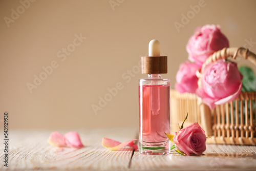 Bottle of essential rose oil and flowers on white wooden table against beige background, space for text photo