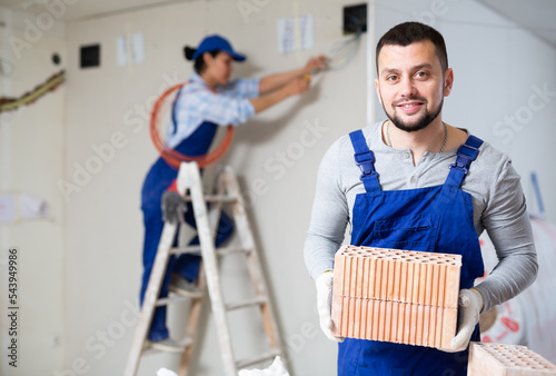 Portrait of skilled confident young bearded builder in blue overall stacking red bricks on construction site indoors, looking at camera with smile