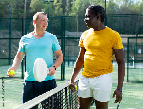 Portrait two sports men of different nationalities, standing on an outdoor court with padel rackets and balls and discussing something © JackF