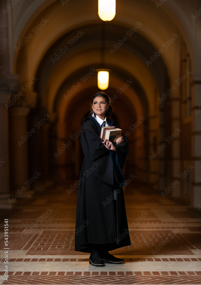 Fototapeta premium School girl in university hall wearing black robe and holding books with magic wand