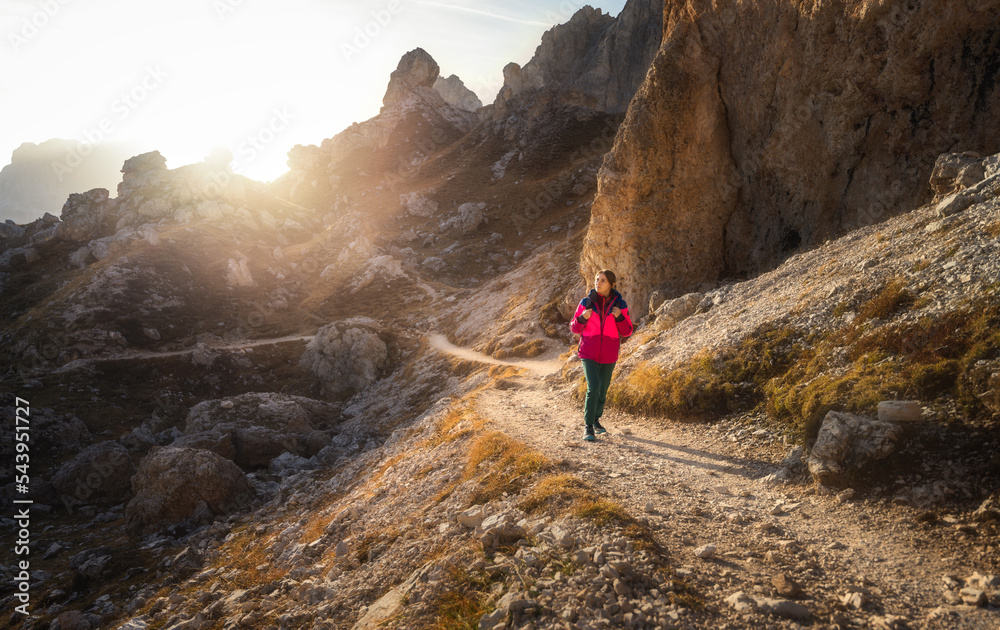 Walking girl with backpack on the trail in mountains at sunset in autumn. Beautiful landscape with young woman, high rocks, path, orange trees, sky in fall in Dolomites, Italy. Adventure and hiking