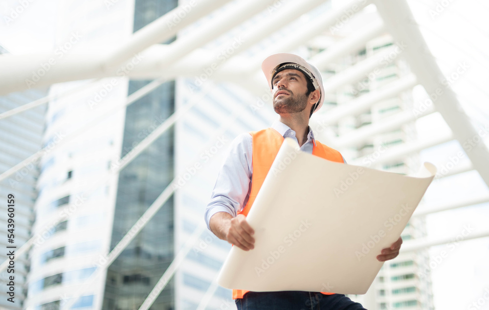 Young caucasian man holding a big paper, a guy wearing a light blue shirt and jeans with an orange vest and white helmet for security in the construction area. Young caucasian man holding a big paper, a guy wearing a light blue shirt and jeans with an orange vest and white helmet for security in the construction area.