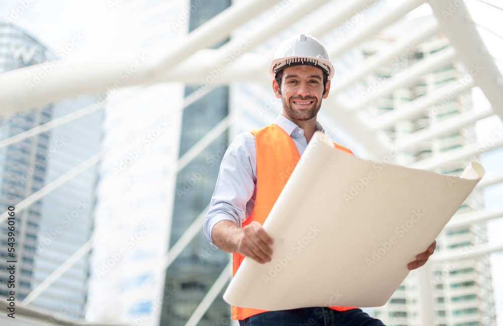 Fototapeta premium Young caucasian man holding a big paper, a guy wearing a light blue shirt and jeans with an orange vest and white helmet for security in the construction area.