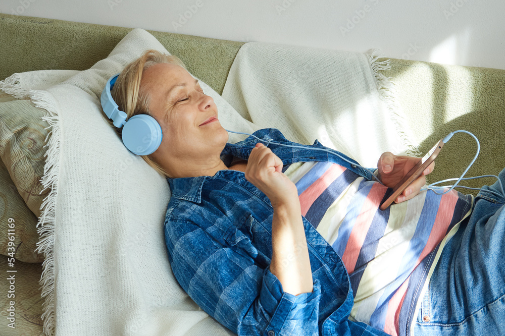 Relaxed mature woman in headphones listens to music, closes her eyes, uses smartphone