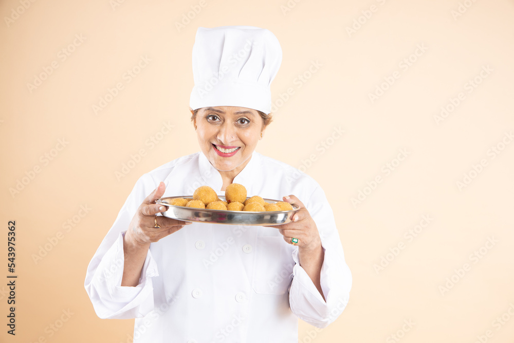 Happy female chef or cook dressed in uniform holding plate full of bundi laddoo or laddus isolated on beige background. Happy female chef or cook dressed in uniform holding plate full of bundi laddoo or laddus isolated on beige background.