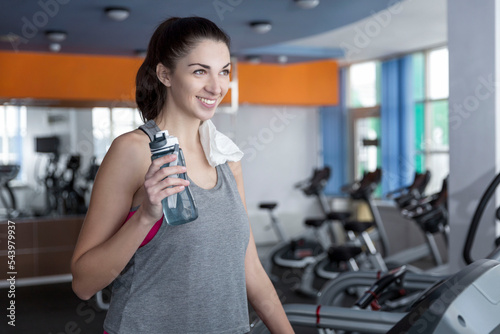 Smiling woman on treadmill with water bottle and towel at the gym. Beautiful young brunette in sportswear. Activity, sport and healthy lifestyle. Vertical.