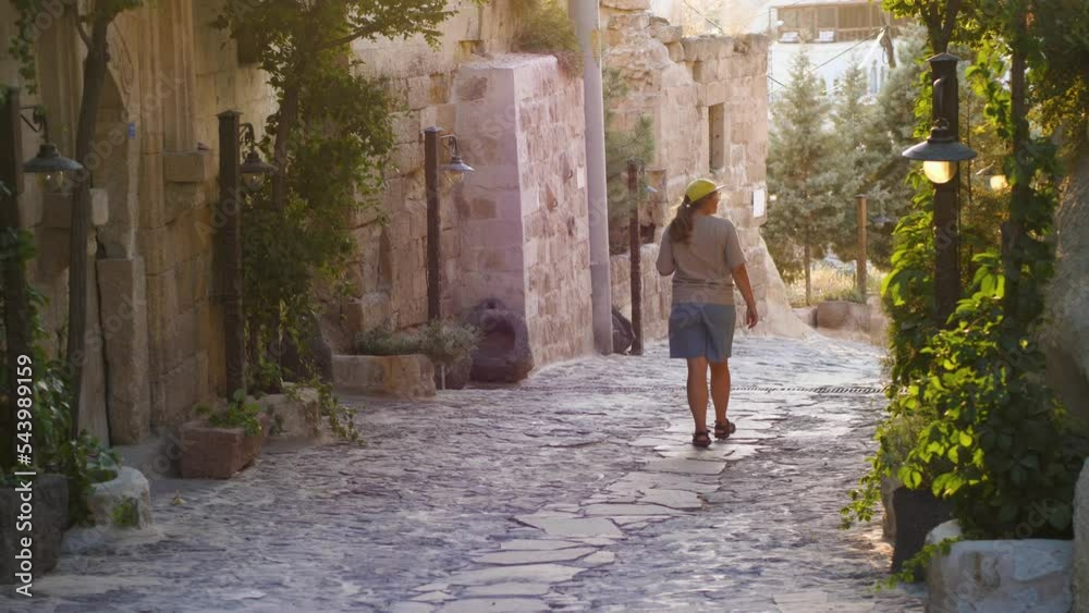 A girl walks through an old street in Cappadocia. A woman walks at sunset for along a stone road among stone houses and street light lanterns. A tourist walks through an old city in Turkey A girl walks through an old street in Cappadocia. A woman walks at sunset for along a stone road among stone houses and street light lanterns. A tourist walks through an old city in Turkey