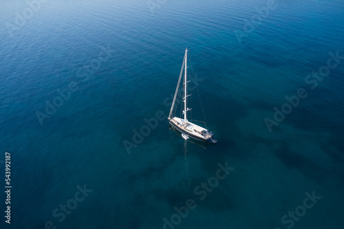 Sailing sports yacht on the water aerial view. A large expensive high-speed sailing yacht is anchored on blue transparent water in the rays of the sun, top view. © Berg