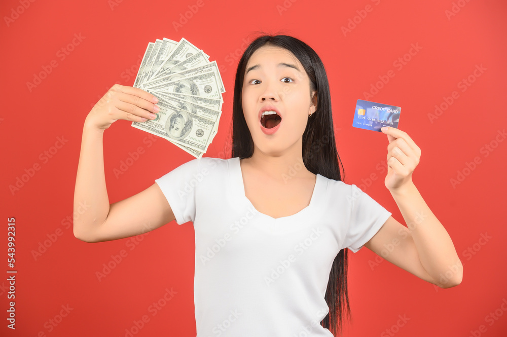 © Johnstocker - Portrait of a cheerful young woman holding money banknotes and credit card isolated over red background