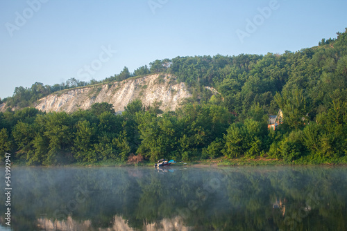 chalk mountain overgrown with dense vegetation, at the foot of the river in the morning haze and a boat near the shore