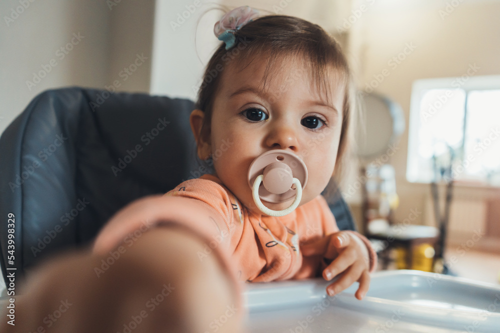 Cute baby girl with a pacifier in her mouth sitting in a high-chair in the kitchen. Space for text. Lifestyle. The baby is having lunch. Funny little gir Cute baby girl with a pacifier in her mouth sitting in a high-chair in the kitchen. Space for text. Lifestyle. The baby is having lunch. Funny little gir