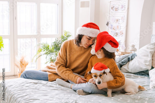 Christmas family portrait on bed in bedroom, baby and dog play, mom and daughter in sweaters and santa claus hat. Have fun in anticipation of Christmas and New Year. Dog jack russell loves his owners