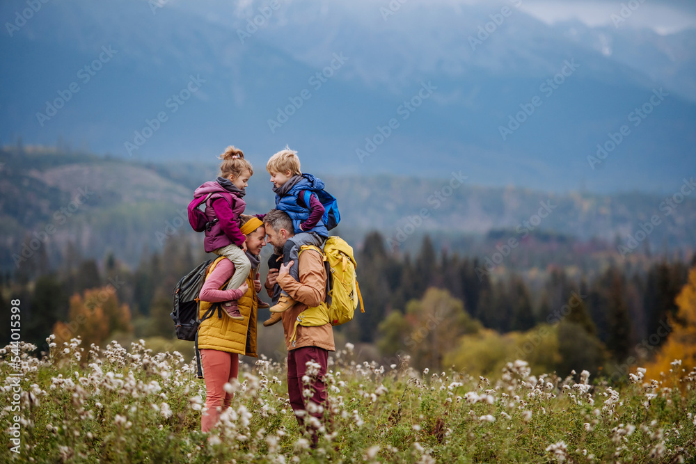 Happy parents with their little kids on piggyback at autumn walk, in the middle of colourfull nature. Concept of healthy lifestyle.