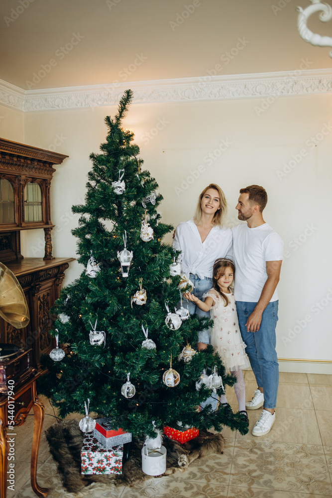 Dad, mom and daughter decorate a Christmas tree. Happy loving family opening gifts. Christmas tree with presents under it. decorated living room. Dad, mom and daughter decorate a Christmas tree. Happy loving family opening gifts. Christmas tree with presents under it. decorated living room.