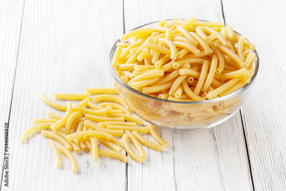 Uncooked maccheroni pasta in glass bowl on white wooden background