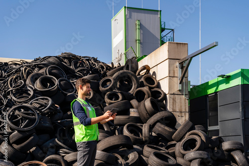 Man with tablet PC standing in front of rubber tires at recycling center photo
