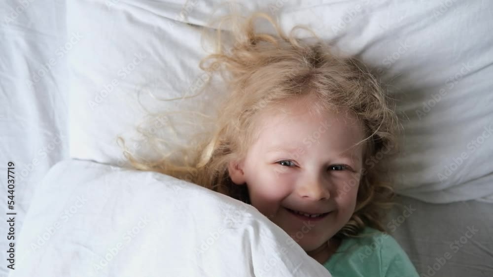 Portrait of a happy Cute funny cheerful little child girl in blue pajamas wakes up in bed in the morning. kid looks at his parents smiling, stretches laughs and hides under the blanket again.
