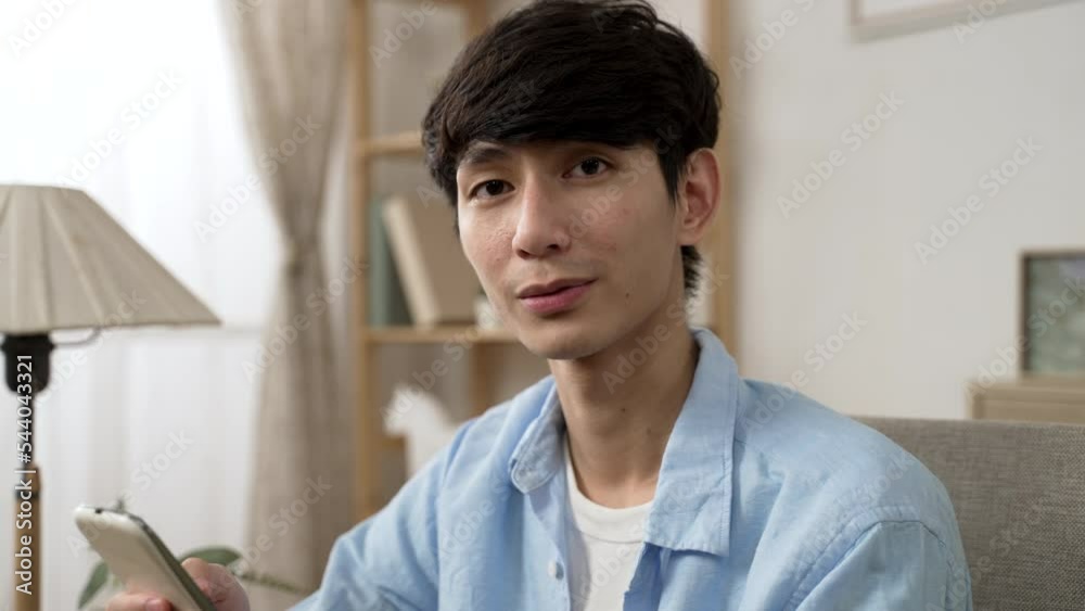 shoulder shot portrait of a cheerful asian man turning to look at the camera with a smile while using his smartphone in the living room at home during daytime.