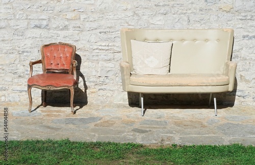 Two chairs in used condition with worn fabric, different styles and padded material.  Empty double seat sofa and armchair near wall with sunlight