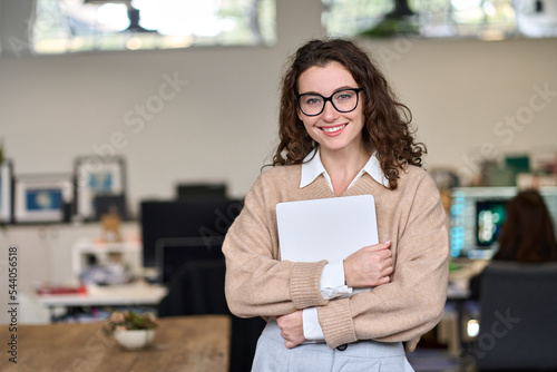 Young smiling professional business woman, happy businesswoman, female company worker intern or corporate manager holding laptop standing in modern office working, looking at camera. Portrait