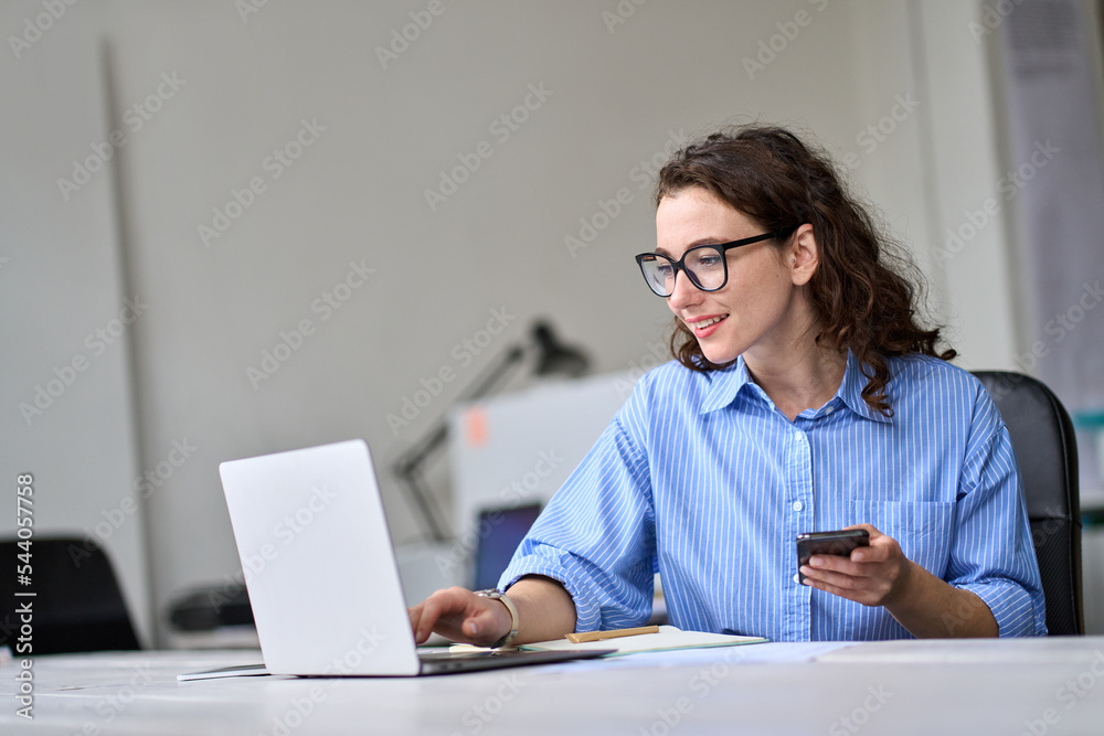 Young business woman company employee worker sitting at desk working online on laptop holding phone. Female professional worker manager using computer mobile technology in corporate modern office. Young business woman company employee worker sitting at desk working online on laptop holding phone. Female professional worker manager using computer mobile technology in corporate modern office.