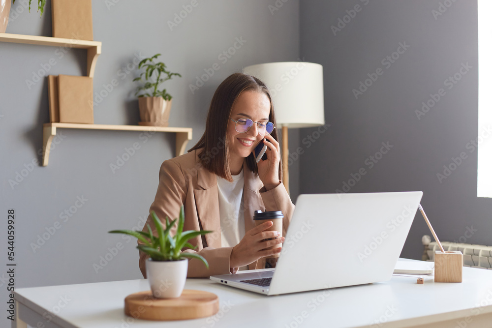 Φωτογραφία Image of beautiful positive attractive young businesswoman working online on notebook in home office and talking on mobile phone, expressing positive emotions, drinking coffee