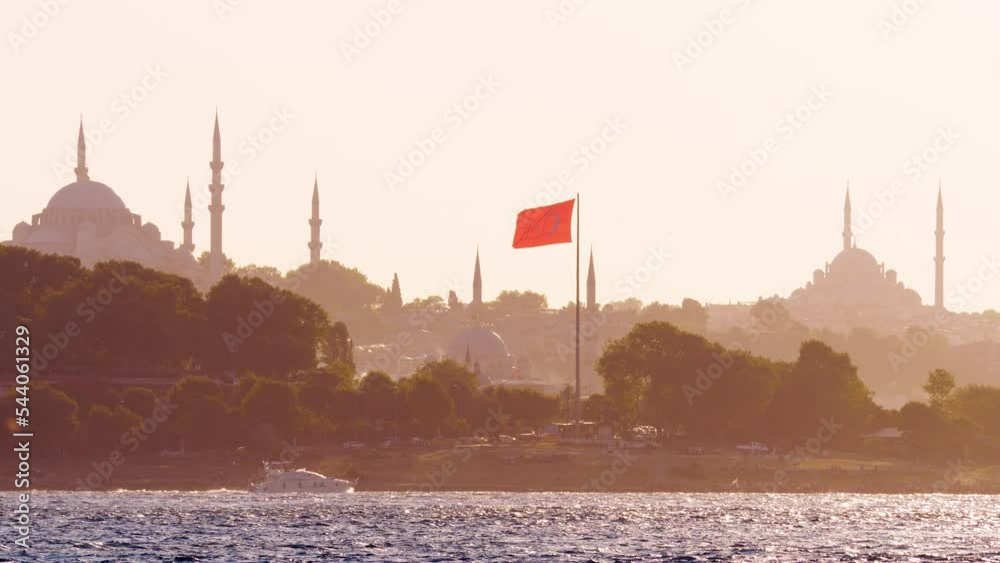 Fatih area of Istanbul seen across Bosporus, telephoto perspective from Asian side. Red flag on tall pole, mosque silhouetted on background. Single yacht slowly cruise on water. Sunny summer evening