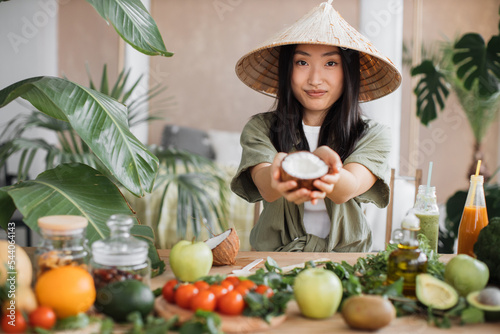 Beautiful asian woman in traditional conical hat holding coconut sitting at table with fruit and vegetables ingredients to make healthy vegan salad, indoor at table at stylish tropical kitchen.
