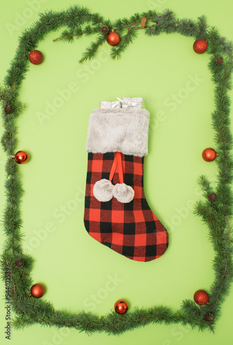 A frame of fir tree branches with red decorations and a Christmas stocking in the center on a pastel green background. Creative Christmas composition. Minimal flat lay.