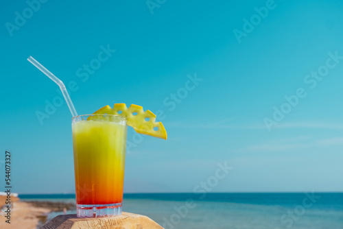 Colorful tropical cocktail in layers on the beach. Decorated with fruit cocktail with straw against the blue sky and the sea.