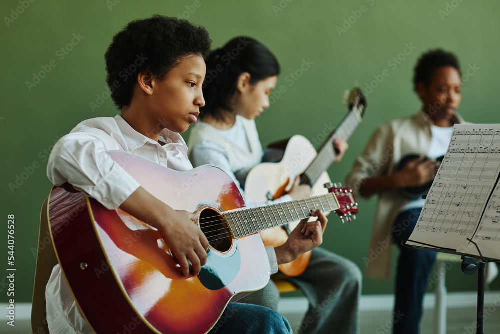 African American schoolgirl with acoustic guitar looking at paper with musical notes while playing instrument against classmates - obrazy, fototapety, plakaty 